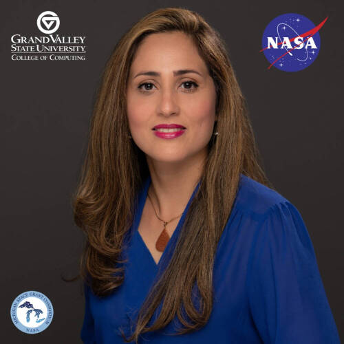 Dr, Sara Sutton wearing a blue chiffon top is staring at a  camera in front of a gray GVSU College of Computing branded cover drop. At the top right, there's a NASA logo. She has long brown hair.
